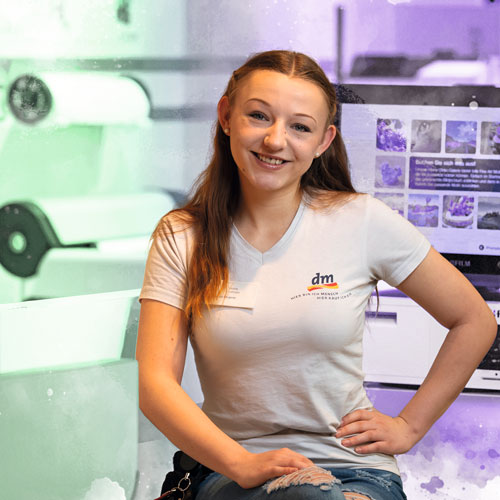 Smiling young woman wearing a white shirt with a dm logo, seated in a bright, lab-like workspace with equipment in the background.