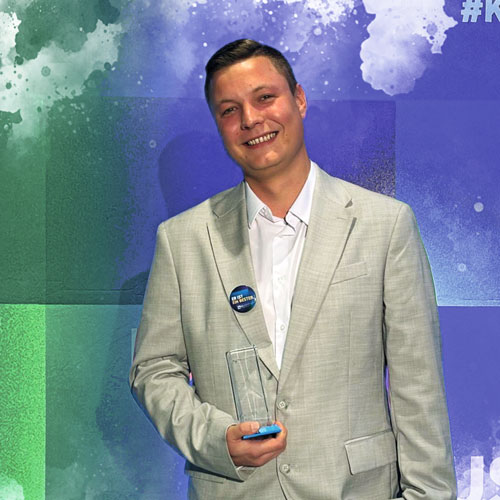 Smiling man in a light gray suit holding a clear glass award and a blue smartphone, against a blue-green abstract backdrop.
