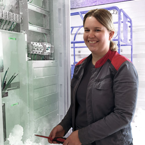 Smiling woman in a grey jacket with red shoulders stands beside an open electrical panel, holding a red tool.