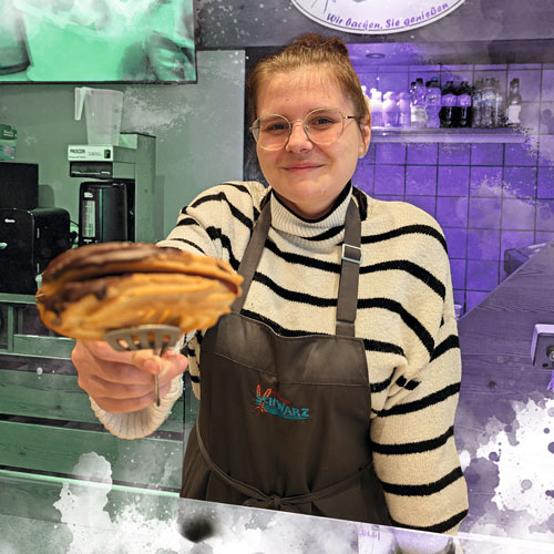 Smiling bakery staff member in a striped sweater and brown apron offers a flaky round pie on tongs toward the camera.