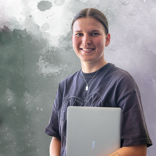 Young person smiling while holding a silver Dell laptop in front of a gray-green abstract background.