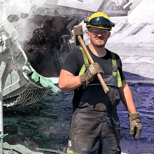 Male worker in safety gear (hard hat with visor, gloves, overalls) holds a wooden-handled axe near a large rock/crusher in a quarry.