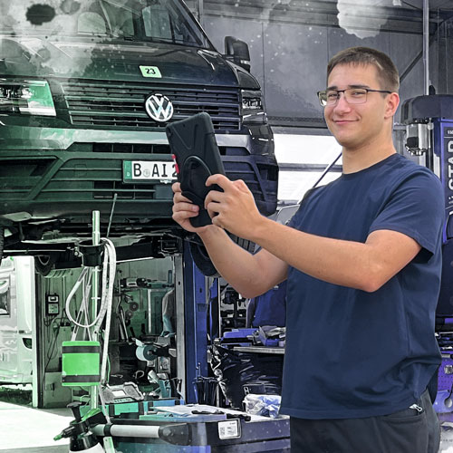 Male mechanic in a blue t-shirt holds a tablet while inspecting a dark blue VW van on a lift in an auto repair shop.