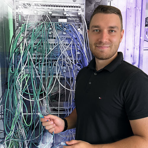 Technician posing beside a network rack filled with tangled blue, green, and gray Ethernet cables.