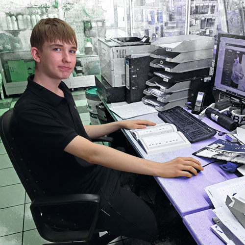 Young man sits at a purple desk in an office, using a keyboard and mouse with printed documents nearby.