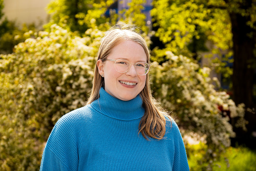 Smiling woman with light hair and glasses wearing a bright blue turtleneck, outdoors with green shrubs in the background.
