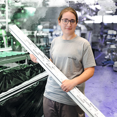 Female factory worker in a gray SCHUCO shirt holding a long measuring rod over a production line.