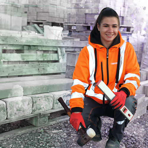 Construction worker in orange safety jacket kneeling with tools beside stacked concrete blocks.