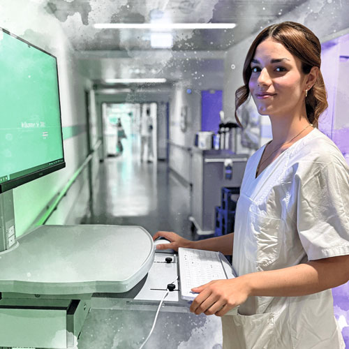 Female healthcare worker in white scrubs standing at a medical console, typing at a keyboard in a hospital corridor.