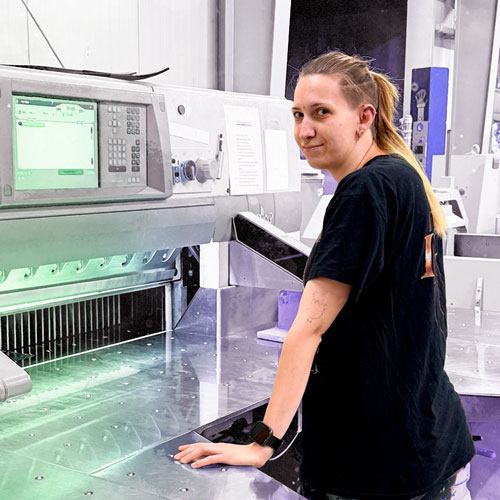 Woman in a black T-shirt stands at a large industrial fabric cutter, hand resting on the metal work surface and looking at the camera.
