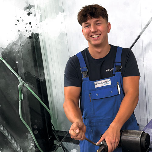 Young man in blue work overalls smiling while operating a lathe in a workshop.